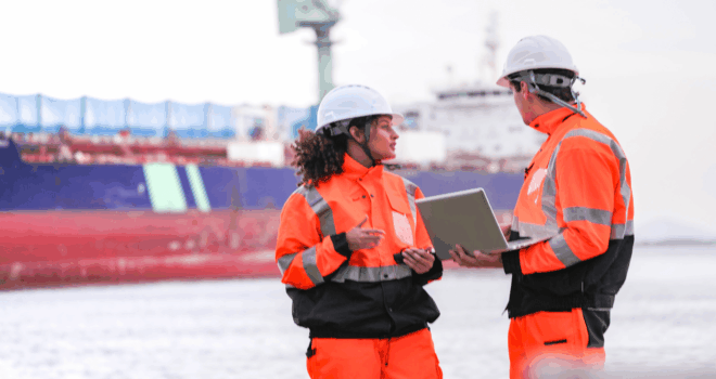 Two port engineers in high-visibility orange uniforms and hard hats communicate while inspecting dockside logistics using a laptop and walkie-talkie, symbolizing teamwork and safety.