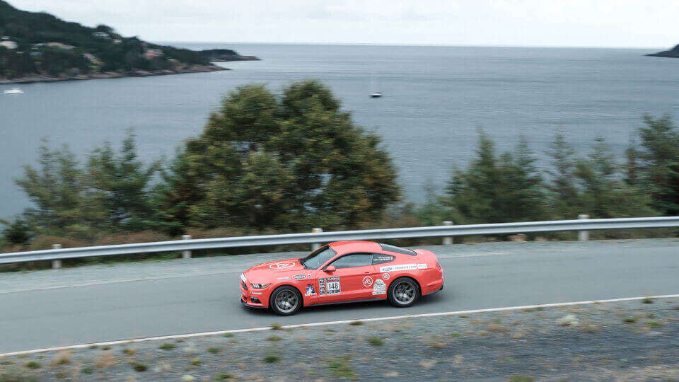 Targa competitor driving on a road with a bay in the background